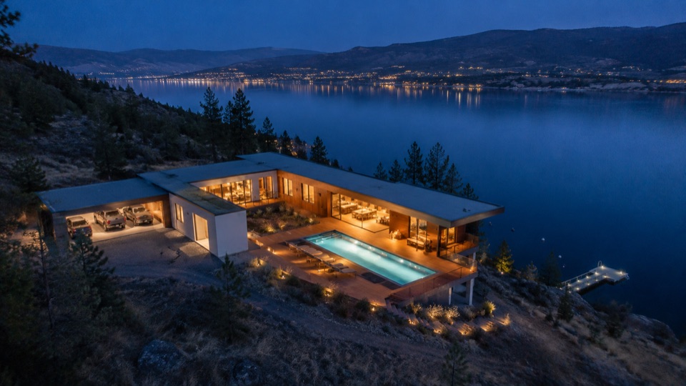 Wider on the same house. The whole hillside, the pool terrace cantilevered, the far shore lit across the water.