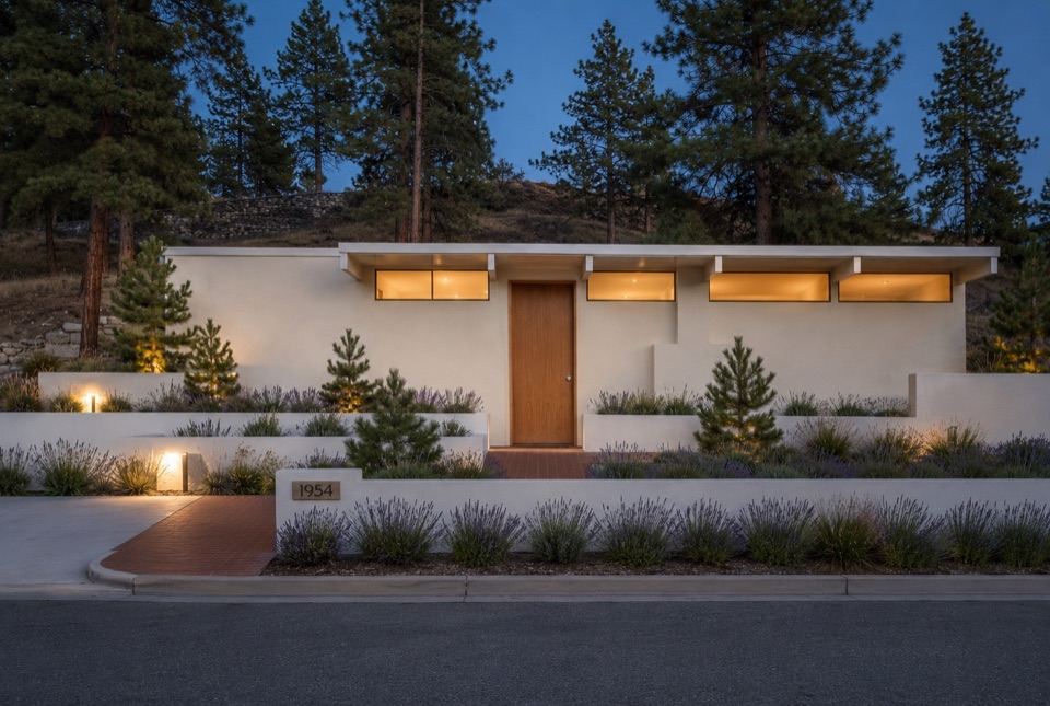 Street elevation at blue hour. A closed stucco face, a slim clerestory band, and the oak door.